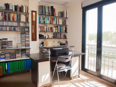 Bright home office featuring a dark wood desk, extensive bookshelves filled with books and CDs, and direct access to an outdoor balcony via a glass door.