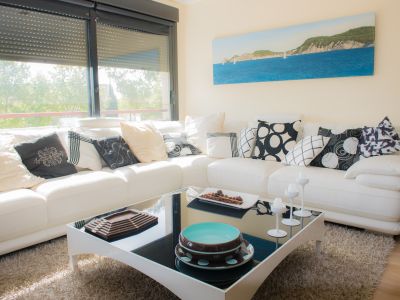 Bright living room: white sectional sofa, patterned cushions, black glass coffee table. Natural light from a window with a dark roller blind. Coastal painting on the wall.