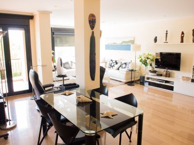 Bright, open-plan living and dining area featuring light wood flooring. A modern glass table with black chairs, a white sofa, and dark-framed glass doors opening to a terrace are visible.