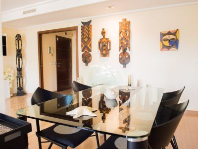 Contemporary dining area featuring a glass table, sleek black chairs, and light wooden flooring. Unique ethnic masks adorn the wall.