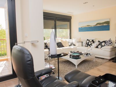 Contemporary living room featuring a white sectional sofa, black glass coffee table, and light wood-look tiled floor. Direct access to a terrace with metal railings and green tree views.