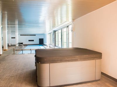 Covered jacuzzi in the foreground, with an indoor heated pool and large windows bringing natural light to this wellness area.
