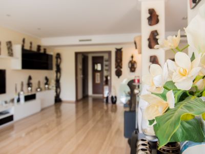 Foreground vase with elegant white and yellow artificial flowers. Bright interior with light wood flooring, leading to a modern living area with decorative wall art.