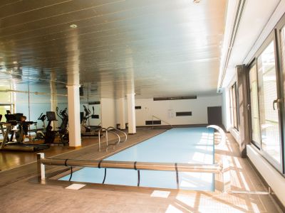Indoor pool with a protective cover, flanked by wooden decking. Large windows flood the space with natural light, and a gym is reflected in the background mirror.