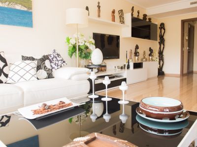 Living room featuring a white sofa, graphic cushions, and a glossy black coffee table with decorative accents. TV unit with shelves and African sculptures.