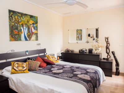 Master bedroom featuring a double bed with a dark headboard and matching nightstands. An abstract painting adorns the wall, complemented by a chest of drawers with mirrors and a ceiling fan. Light wood-look flooring.