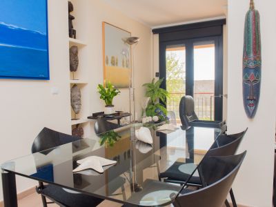 Spacious dining area featuring a glass table and black chairs, accented by a blue abstract painting and tribal masks on shelves. Large glass doors open to a balcony, providing ample natural light.