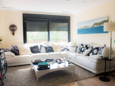 Spacious living room featuring a white sectional sofa, modern glass coffee table, and large window with roller shutters, providing natural light and green views. Ducted air conditioning visible.