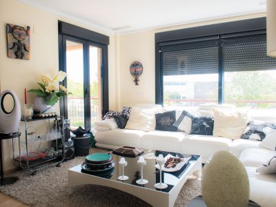Spacious living room featuring a white sofa with diverse cushions, a plush rug, and a modern coffee table. Large windows with black frames offer views of outdoor greenery.