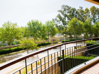 View from a private terrace showcasing lush communal gardens with mature trees, a winding path, and a stone wall, under a bright sky. The balcony features a classic metal railing and terracotta-style tiles.