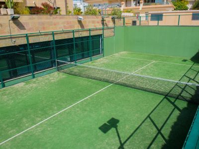 View of the community's outdoor padel court, featuring a vibrant green artificial turf surface, net, and sturdy green mesh walls, with residential buildings visible in the sunny background.