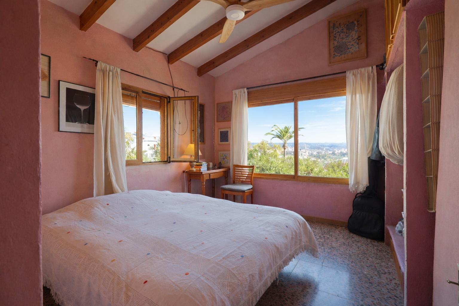 Bedroom featuring exposed wooden beams, warm-toned walls, and large windows offering views of the surrounding Mediterranean landscape and palm trees.