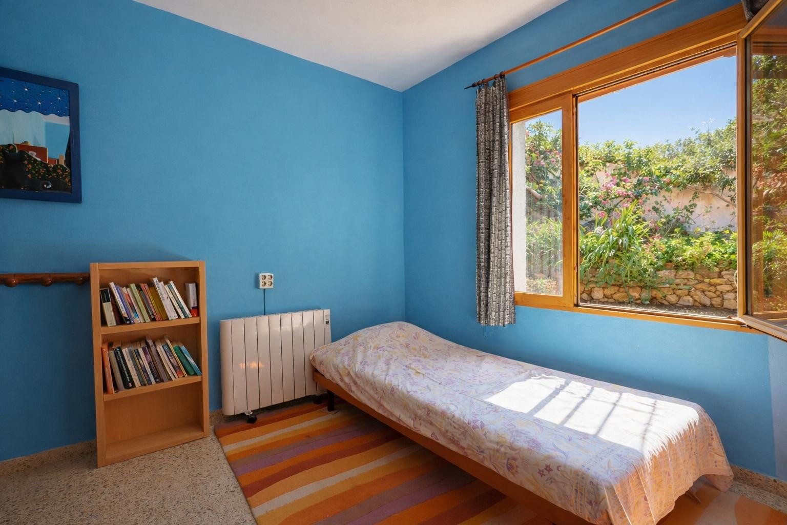 Bedroom featuring vibrant blue walls, a single bed, and a wooden bookshelf. A large wooden-framed window provides natural light and views of the garden's stone walls.