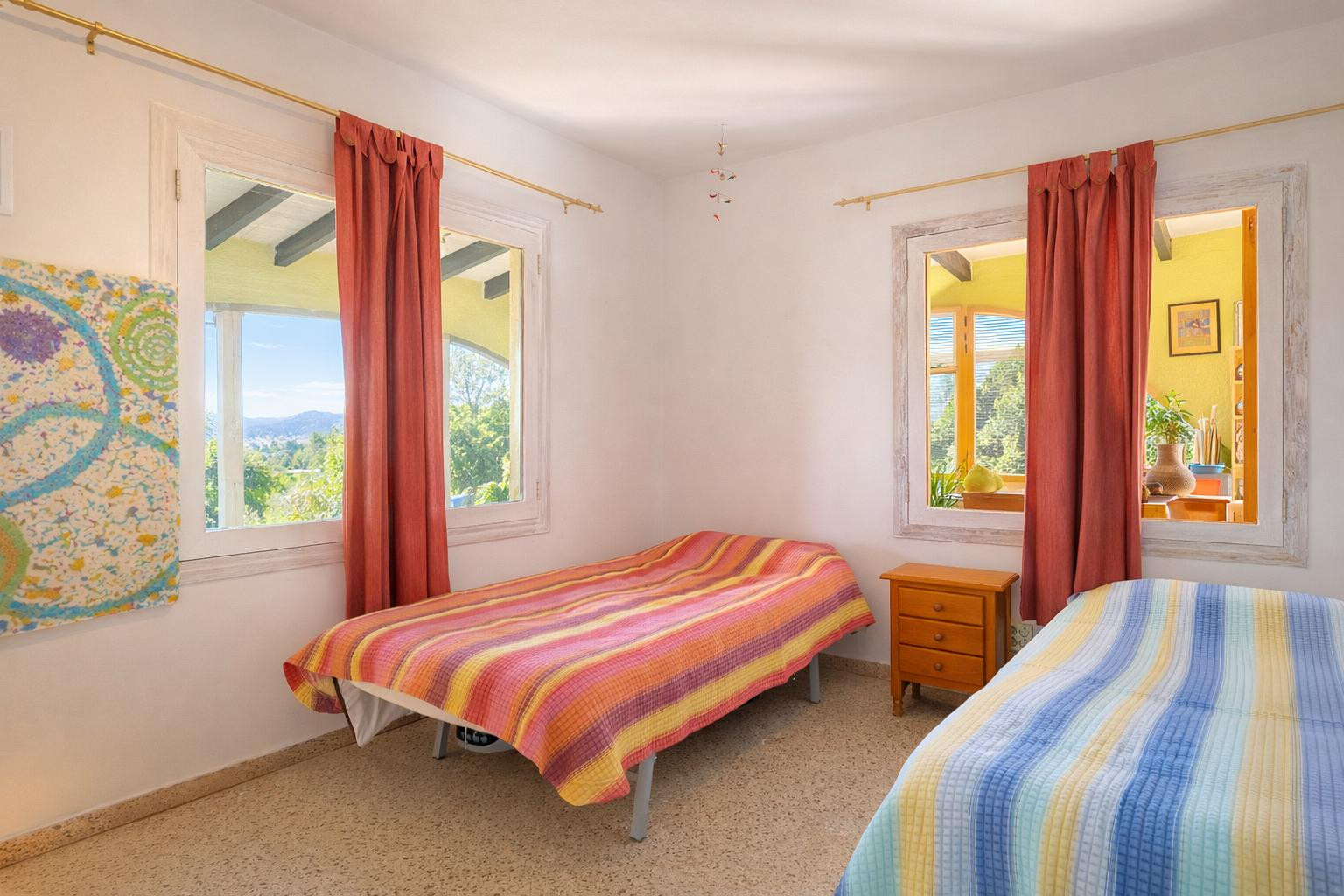 Bright bedroom featuring twin beds, a wooden nightstand, and dual-aspect windows with white frames overlooking the natural landscape and interior spaces.
