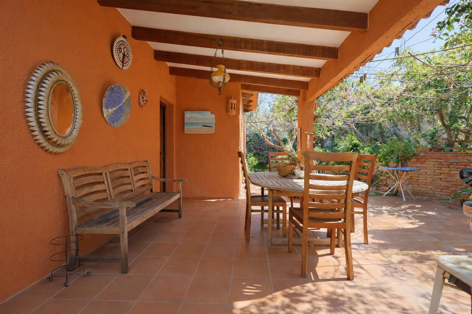 Covered terrace featuring exposed wooden beams, terracotta flooring, and a wooden dining set against warm ochre walls.
