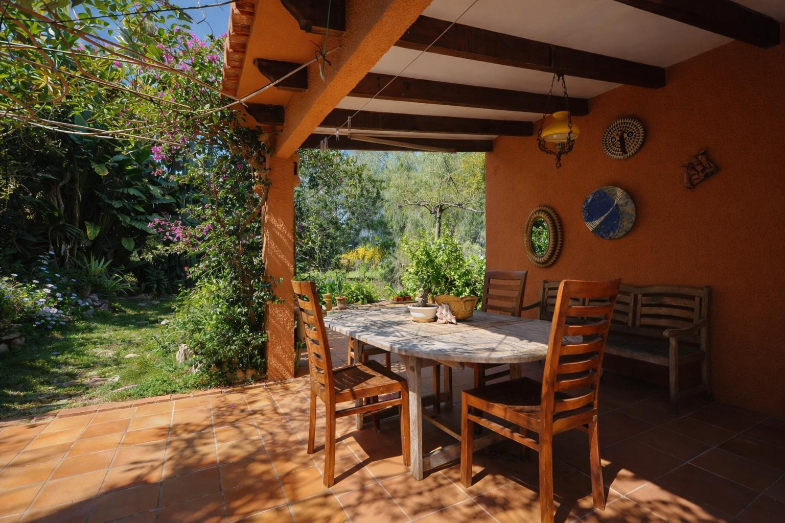 Covered terrace featuring exposed wooden beams, terracotta flooring, and an outdoor dining set overlooking a lush Mediterranean garden.