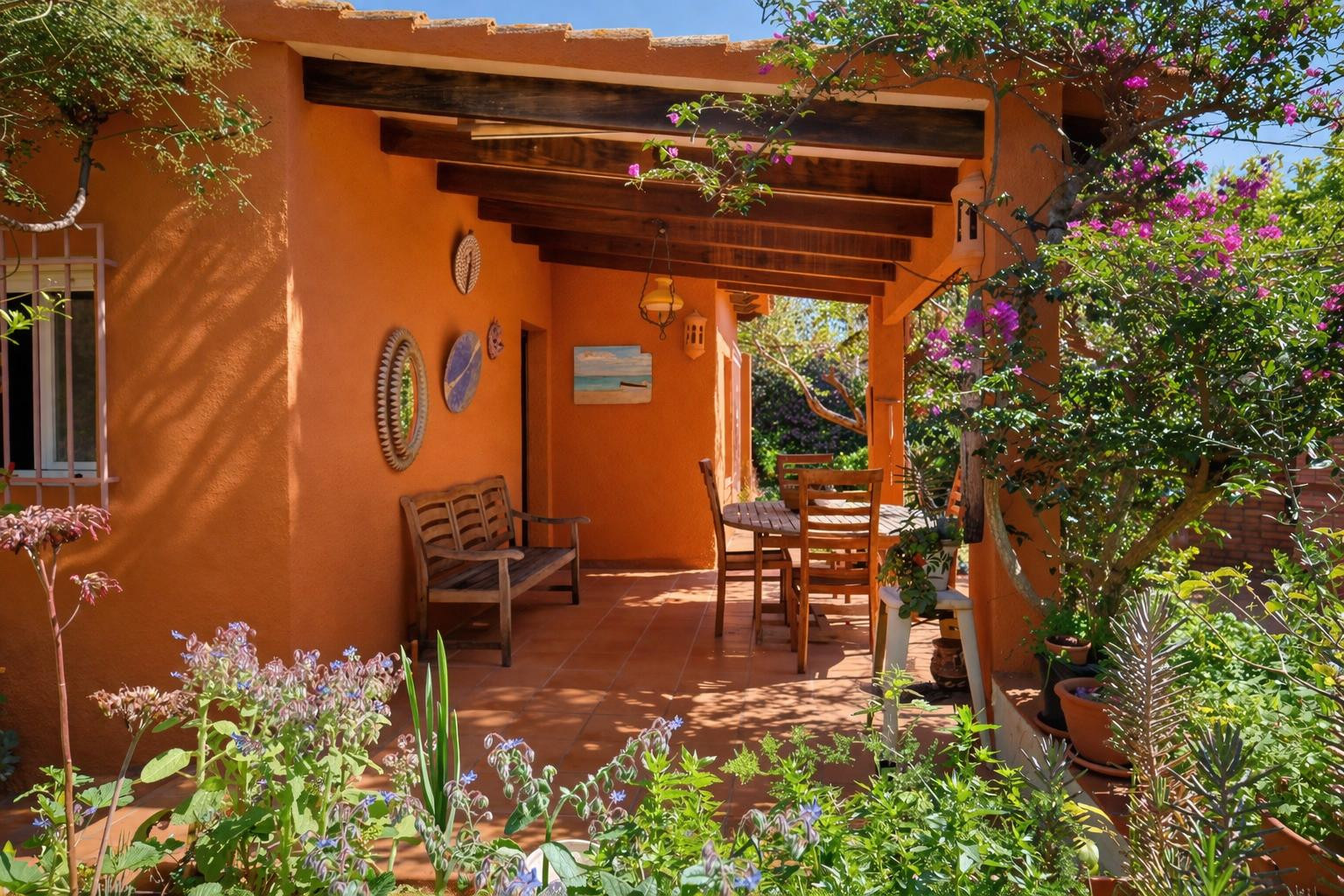 Covered terrace featuring exposed wooden beams, terracotta flooring, and rustic wooden furniture, framed by lush Mediterranean gardens.