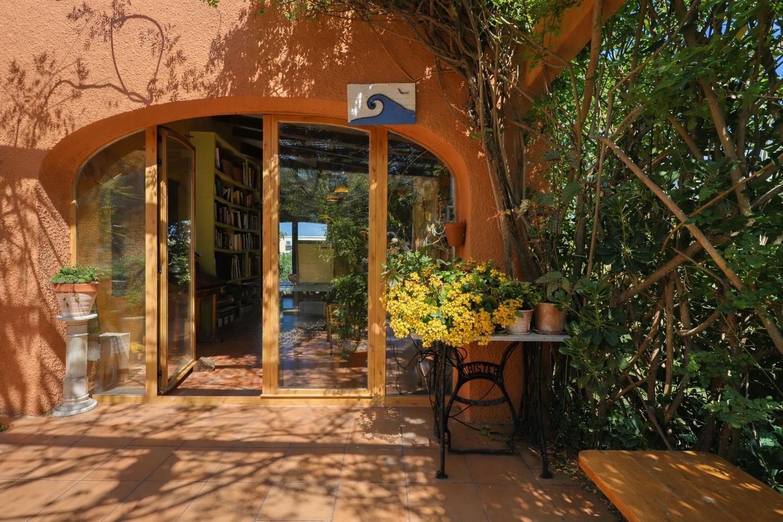 Exterior entrance with arched wooden frames and glass doors leading to a library. Features terracotta flooring and lush foliage creating natural dappled light.