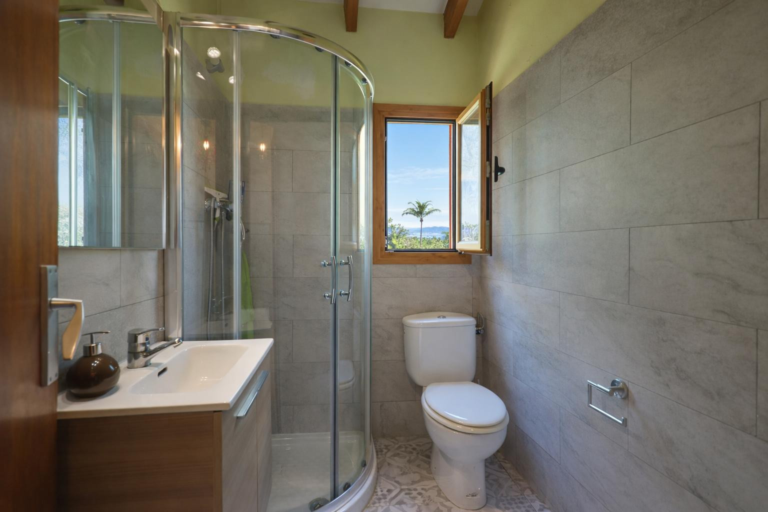 Functional bathroom featuring a curved glass shower, grey tiling, and wooden ceiling beams. A window provides natural light and views of the surrounding landscape.