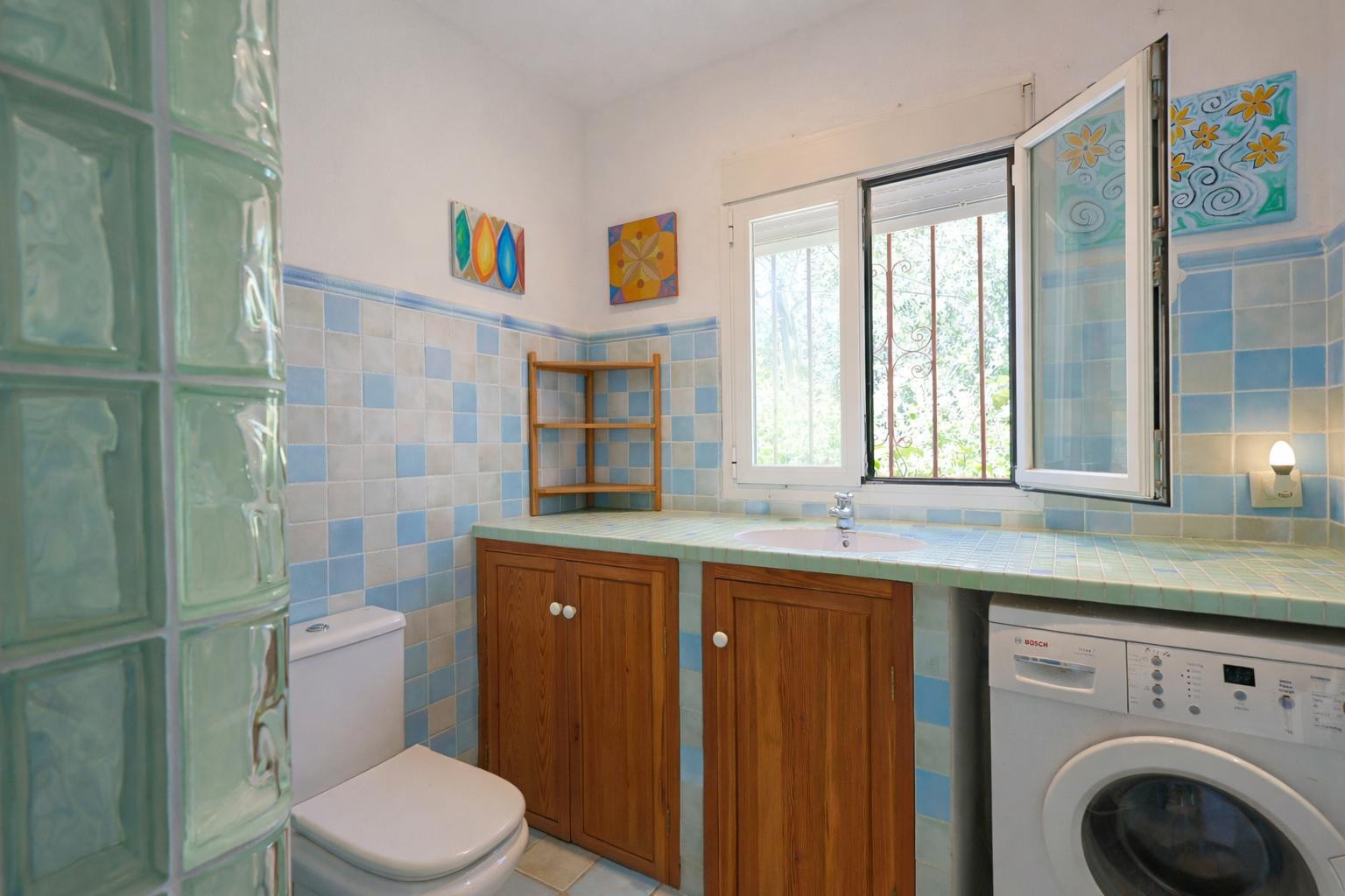 Functional bathroom featuring blue-toned tiling, a tiled countertop with integrated sink, wooden cabinetry, and a laundry area with natural light.