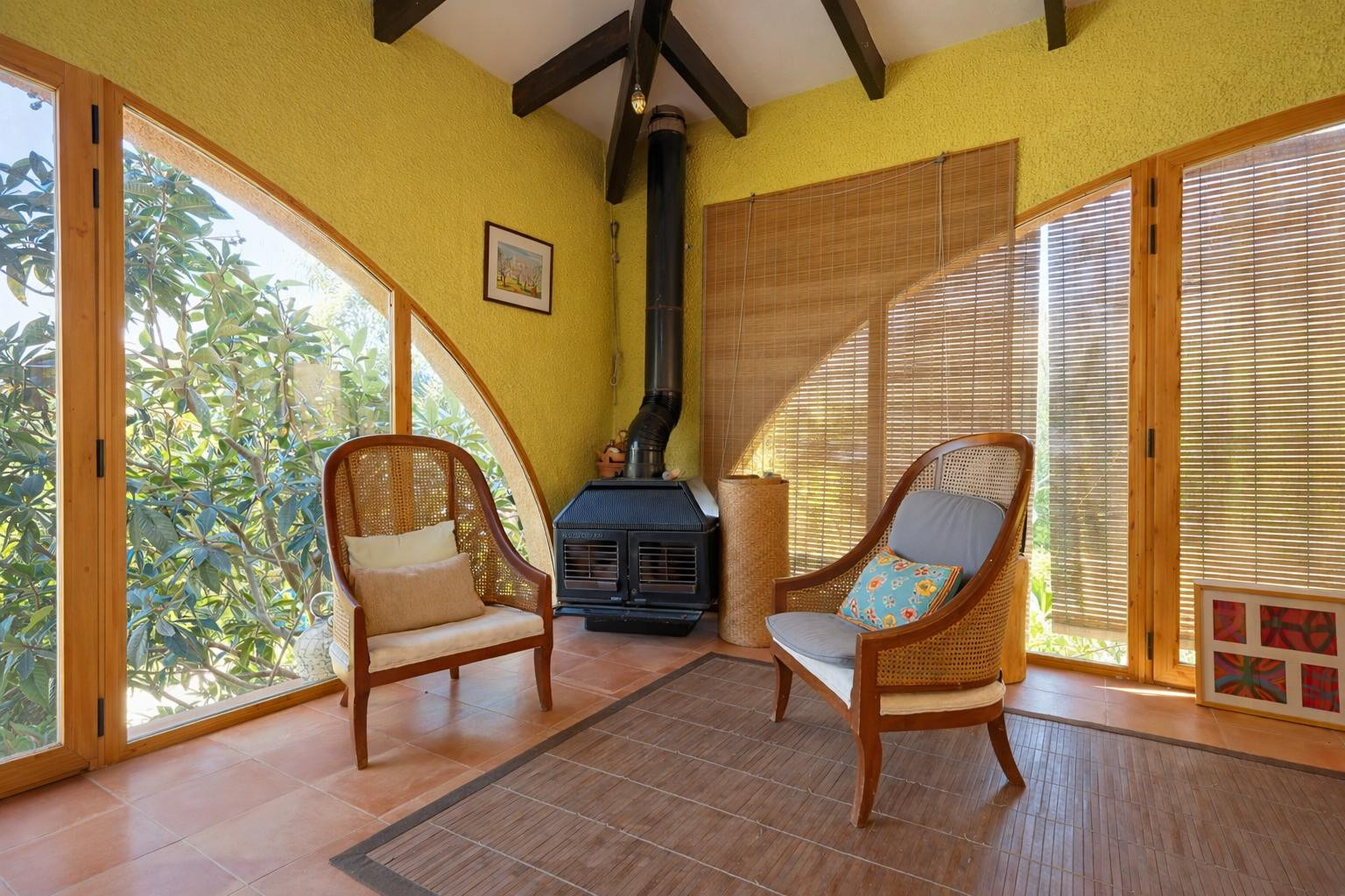 Glazed naya featuring dark wood ceiling beams, a corner pellet stove, and large arched windows with bamboo blinds and terracotta flooring.