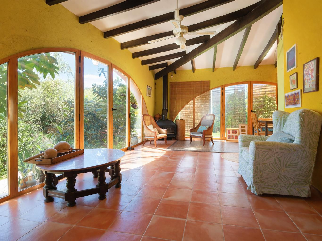 Glazed naya featuring dark wooden ceiling beams, terracotta flooring, and a pellet stove, integrated with the garden through large arched windows.