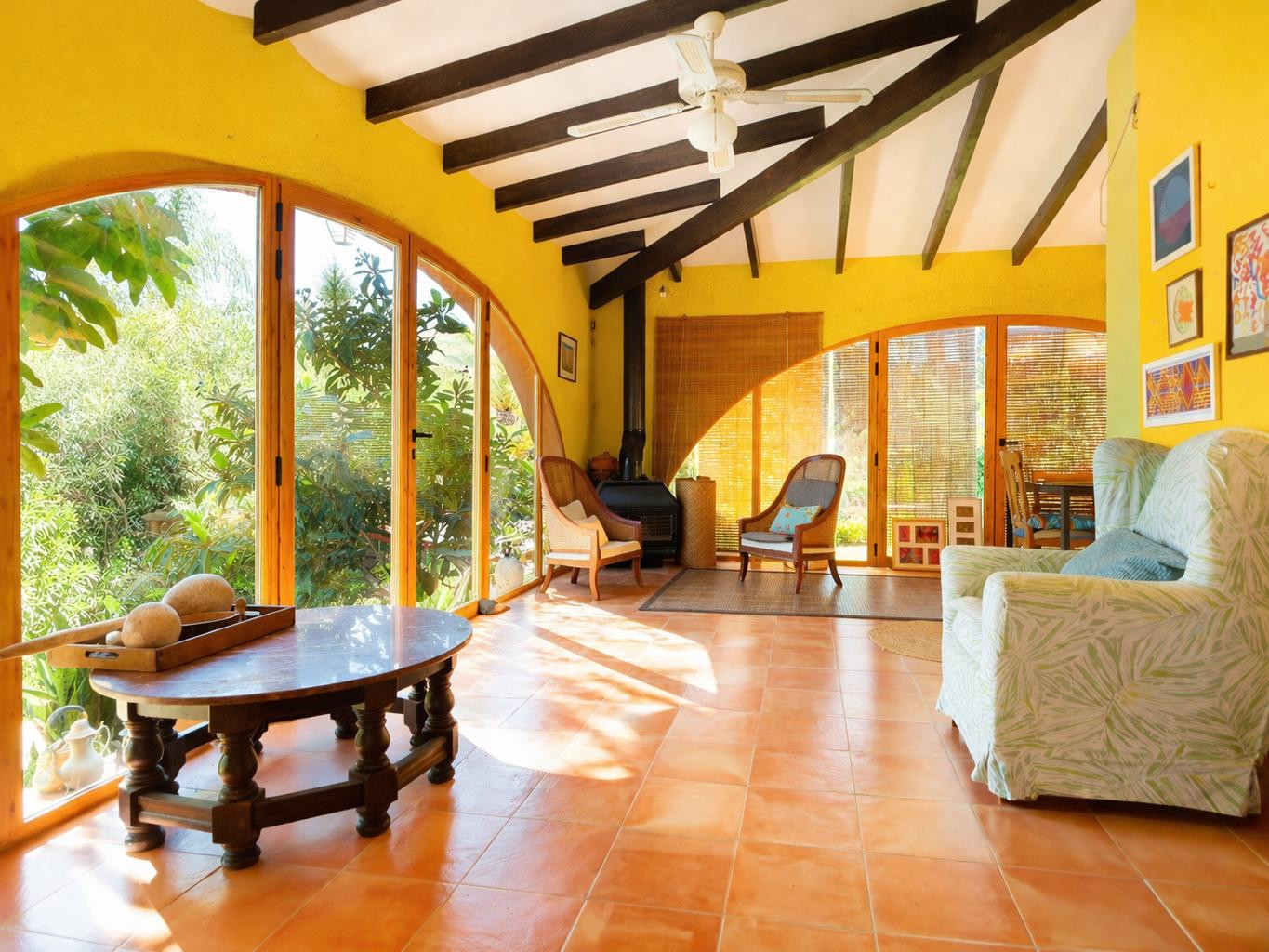 Glazed naya featuring exposed wooden beams, terracotta flooring, and a pellet stove, with large arched windows overlooking the Mediterranean garden.