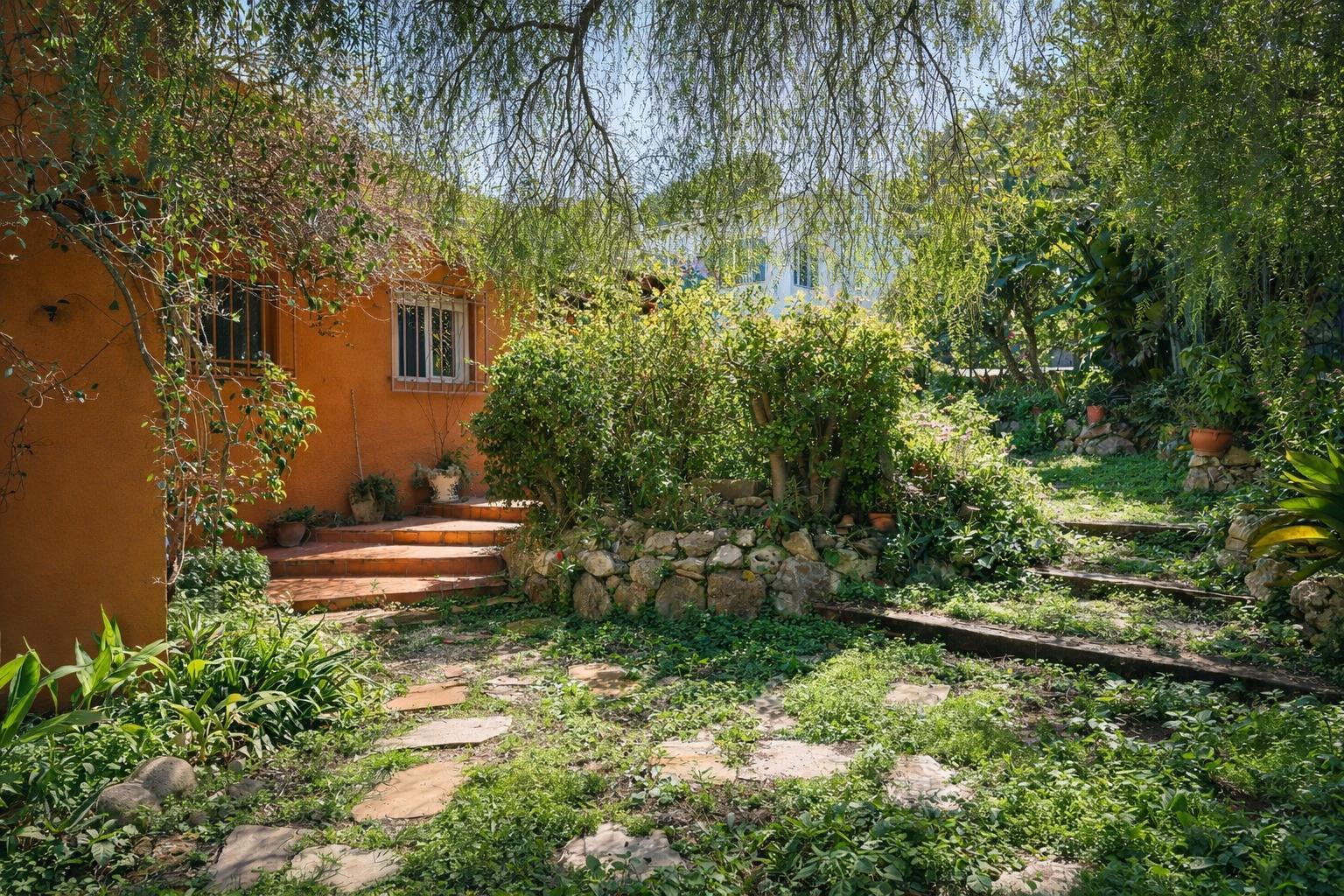 Mediterranean garden featuring stone pathways and wooden sleeper steps, surrounded by mature greenery and natural stone walls against the terracotta facade.