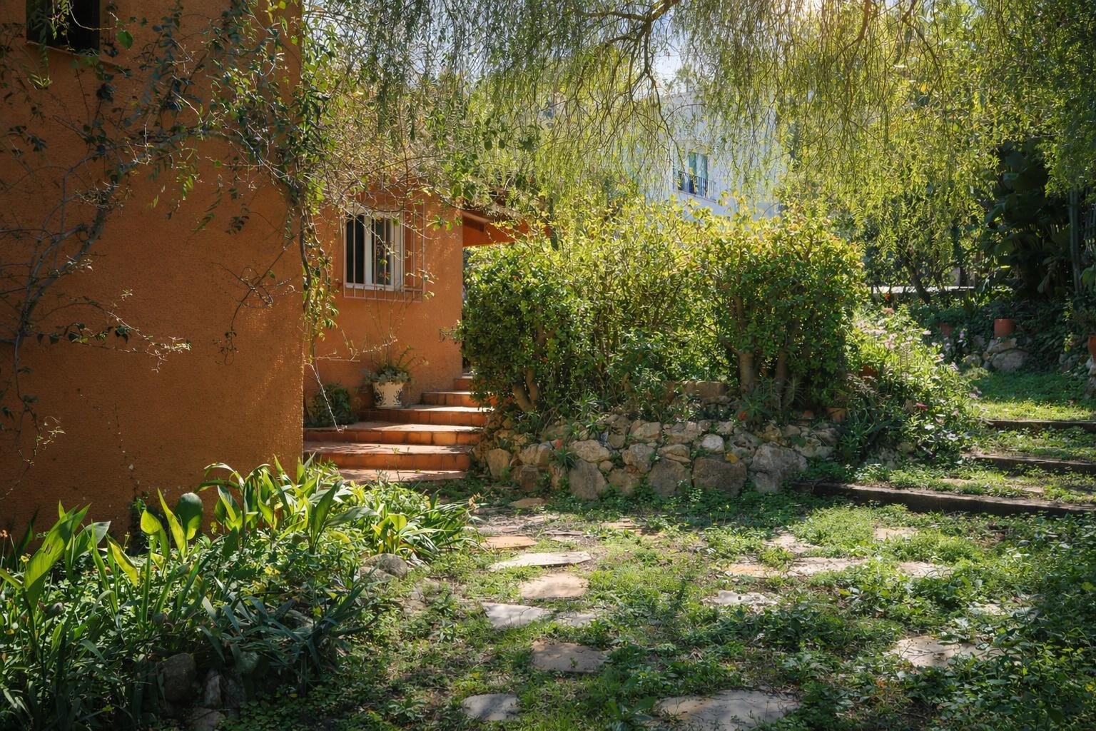 Natural garden featuring stone pathways and terracotta steps leading to the villa's ochre facade, surrounded by mature Mediterranean vegetation.