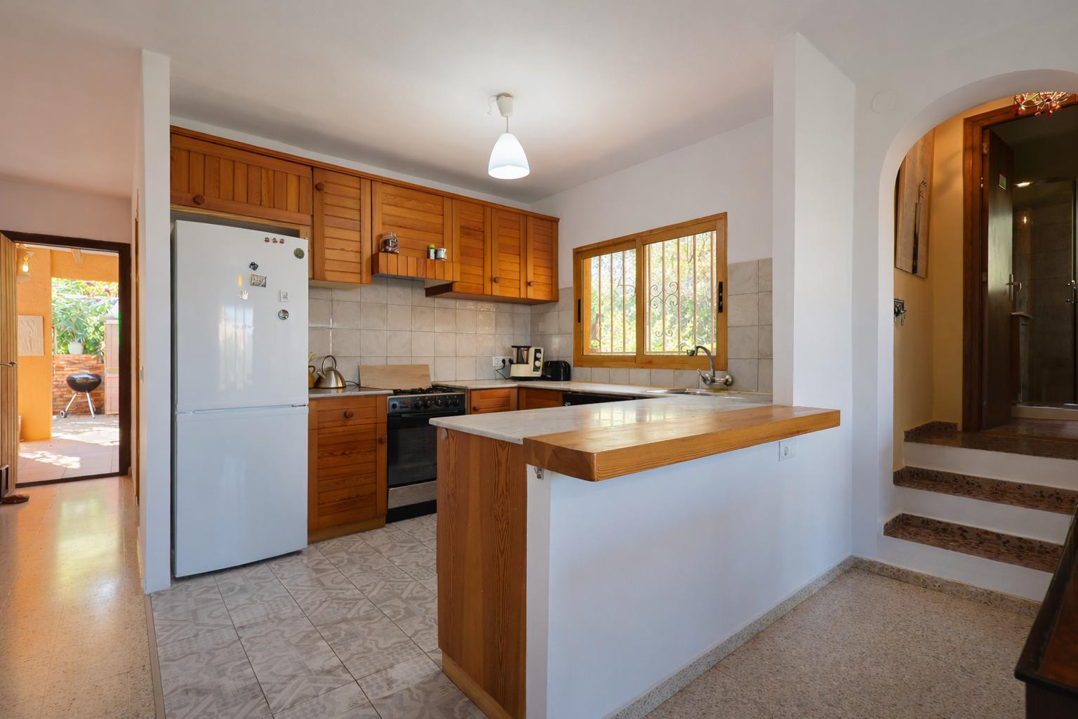 Open-concept kitchen with wooden cabinetry, marble countertops, and a breakfast bar. Features ceramic flooring and a view through to the outdoor terrace and BBQ area.