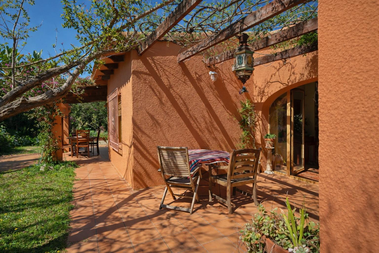 Outdoor terrace featuring terracotta tiling, a wooden pergola with climbing vines, and rustic wooden seating areas against a warm-toned facade.