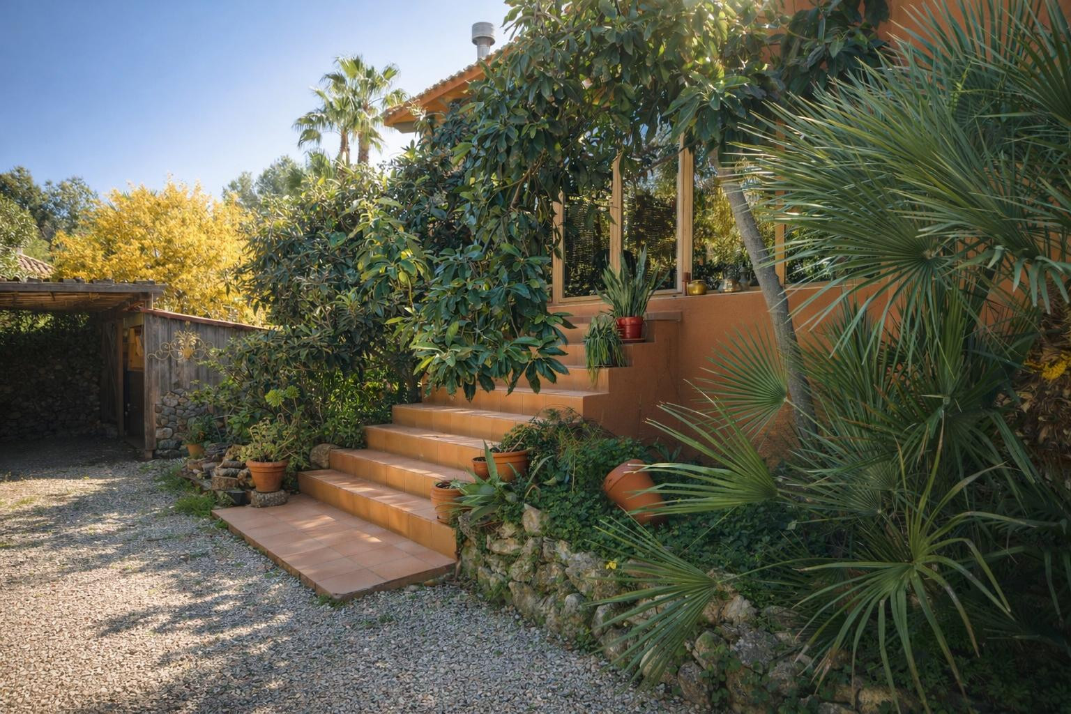 Tiled exterior staircase leading to the glazed porch, featuring lush Mediterranean plants, stone retaining walls, and a gravel driveway area.