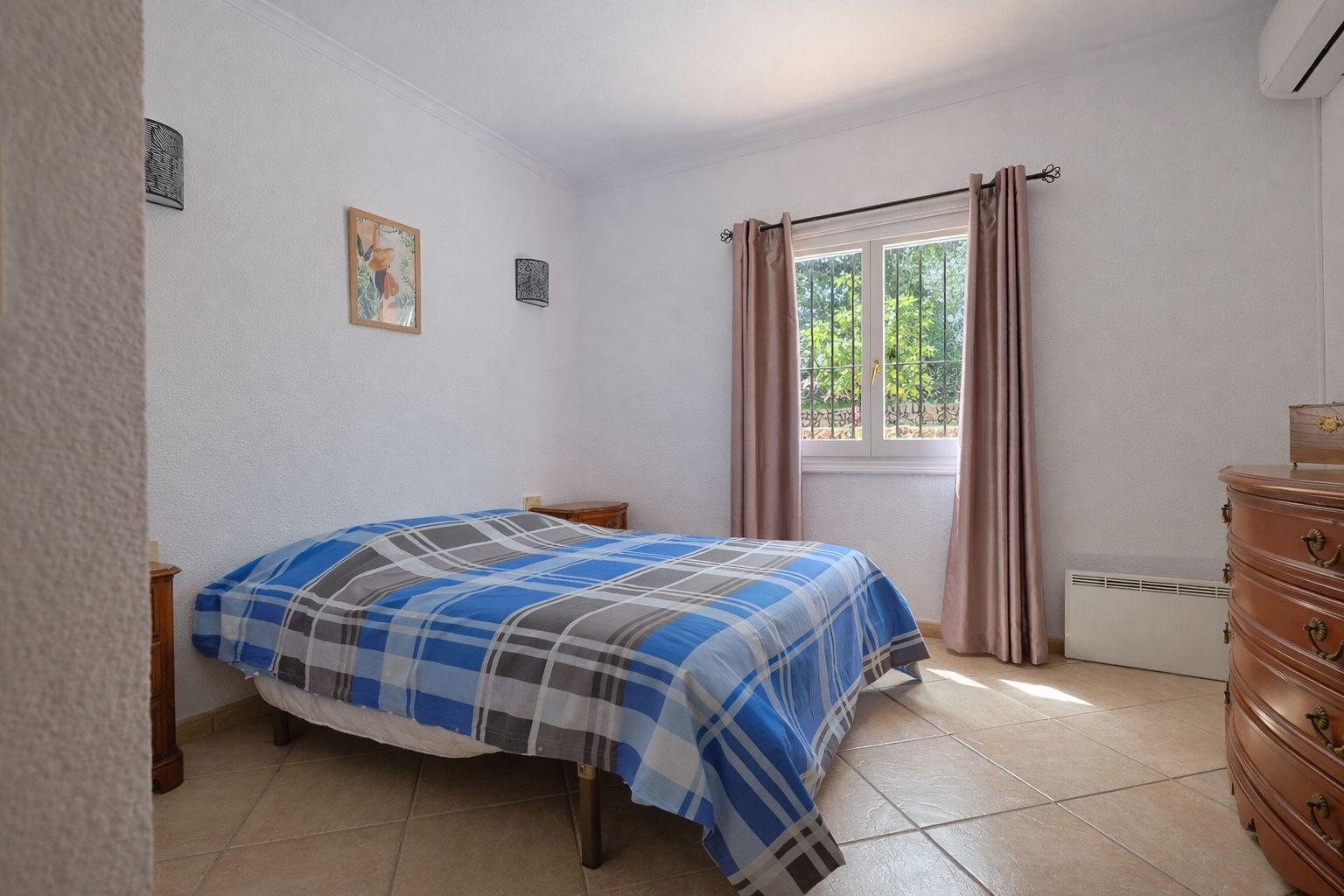 Bright bedroom featuring ceramic tile flooring, classic wooden furniture, and garden views. Includes air conditioning and electric wall heating.