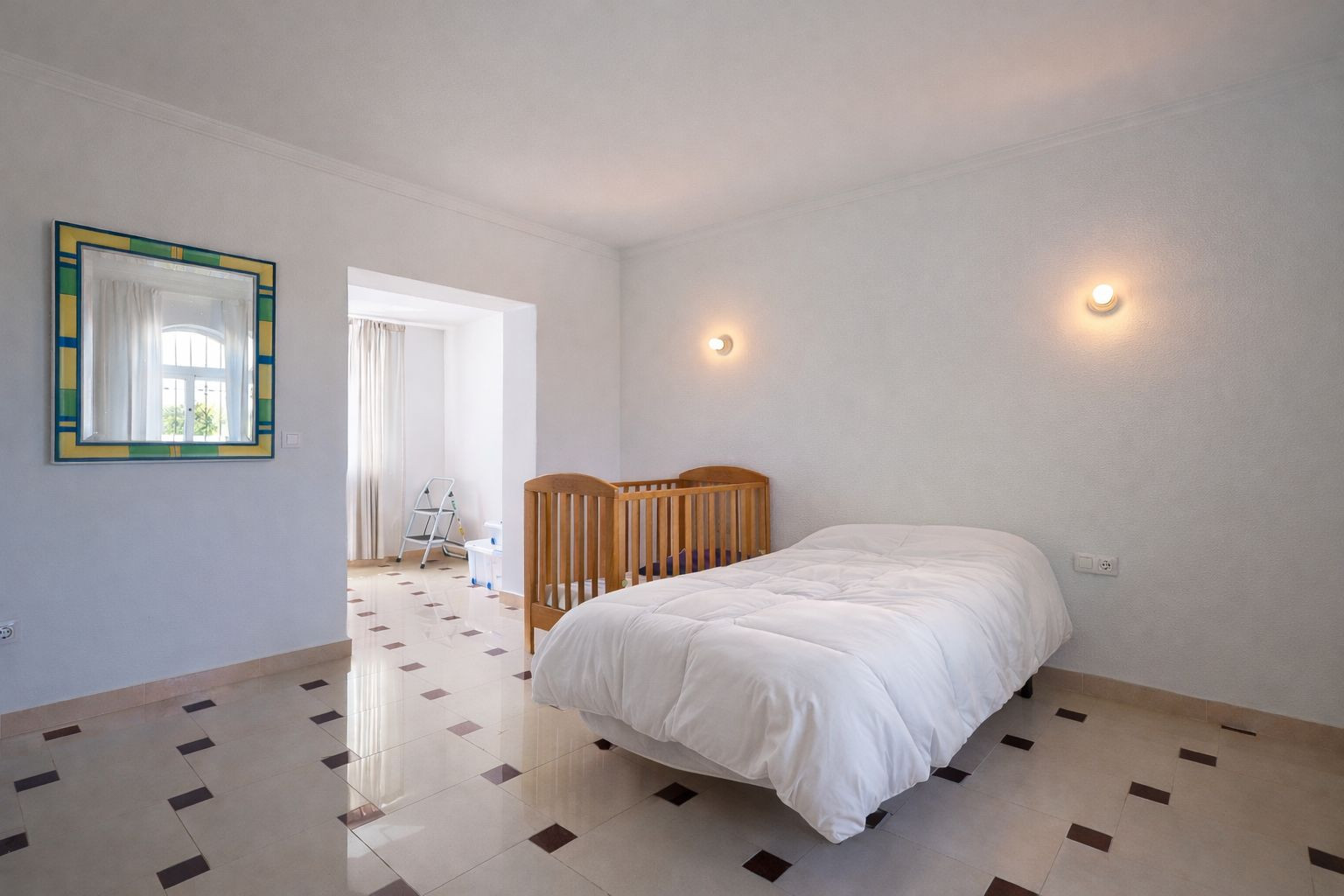 Bright bedroom featuring tiled flooring with dark accents, a single bed, and a wooden crib. Includes wall-mounted sconces and an open doorway to a sunlit area.