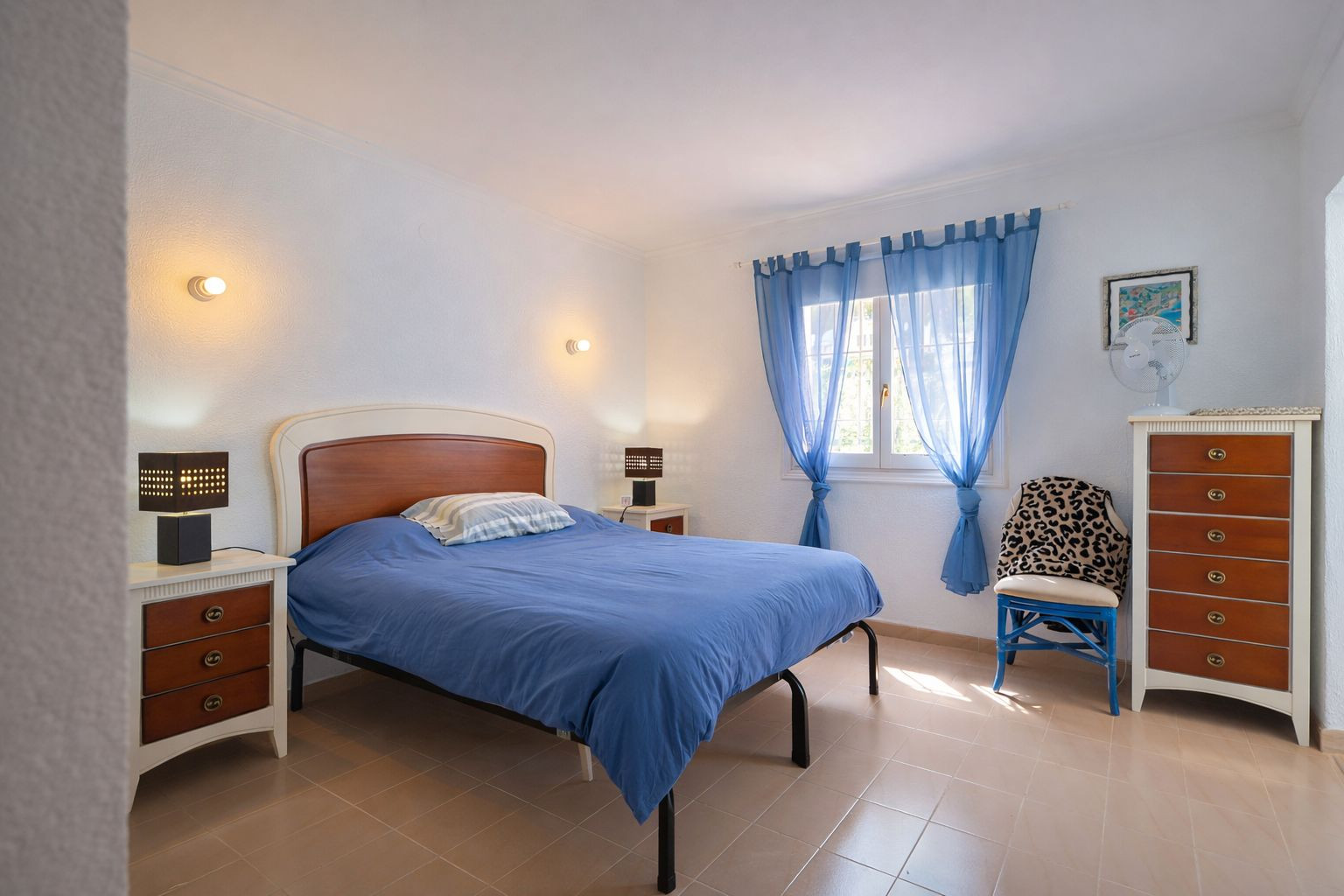 Bright bedroom featuring tiled floors, a double bed with a wooden headboard, and matching furniture. Includes a tall chest of drawers and natural light from the window.