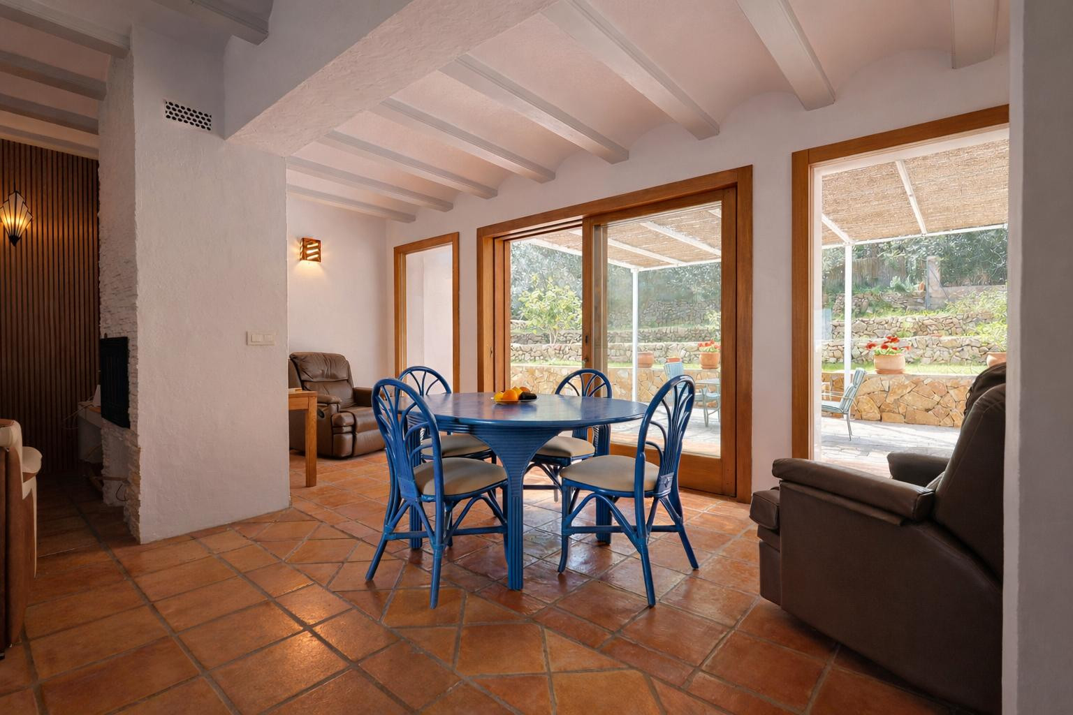 Bright dining area featuring a blue table set, white ceiling beams, and terracotta flooring. Large wooden sliding doors provide direct access to the garden.