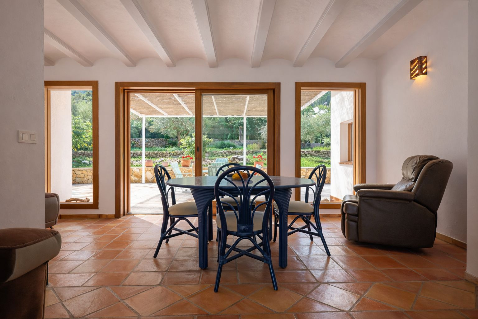 Bright dining area featuring white exposed ceiling beams, terracotta tile flooring, and wooden-framed sliding doors opening onto the covered terrace.