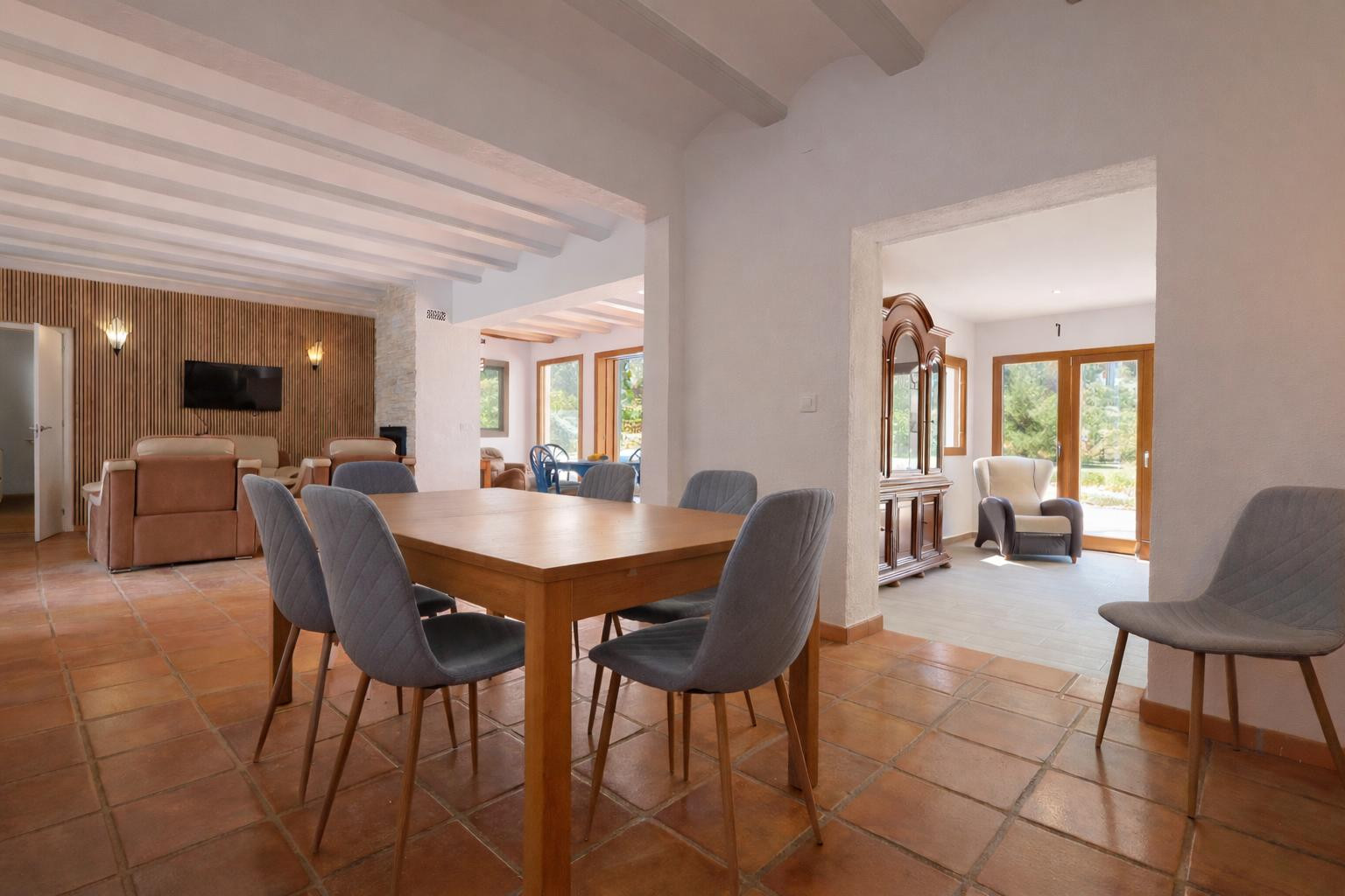 Dining area featuring a wooden table and upholstered chairs, terracotta tile flooring, and white exposed ceiling beams connecting to the various living spaces.