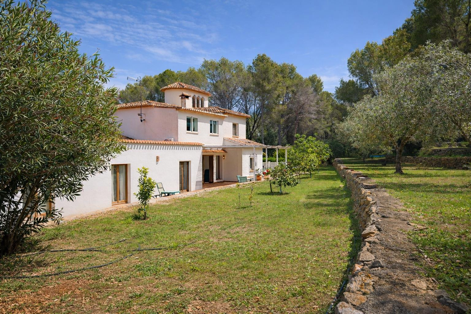 Exterior view of the white-facade villa featuring a terracotta tile roof, set within a garden with young fruit trees and a traditional stone wall.