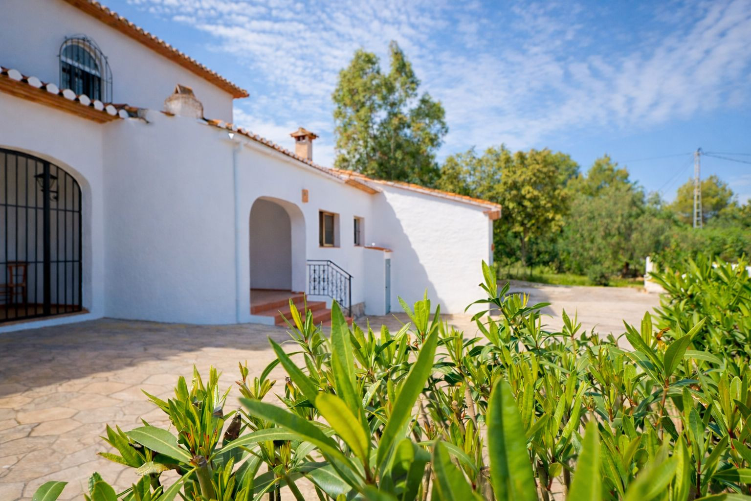 Exterior view of the white-walled villa featuring terracotta roof tiles, a natural stone patio, and arched architectural details surrounded by lush greenery.