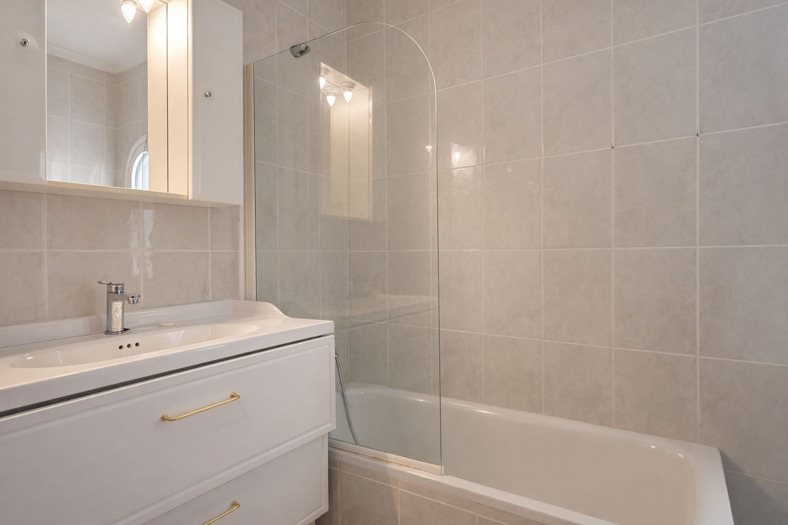 Full bathroom featuring a bathtub with a curved glass screen, white vanity with gold-tone handles, and a mirrored cabinet with integrated lighting.