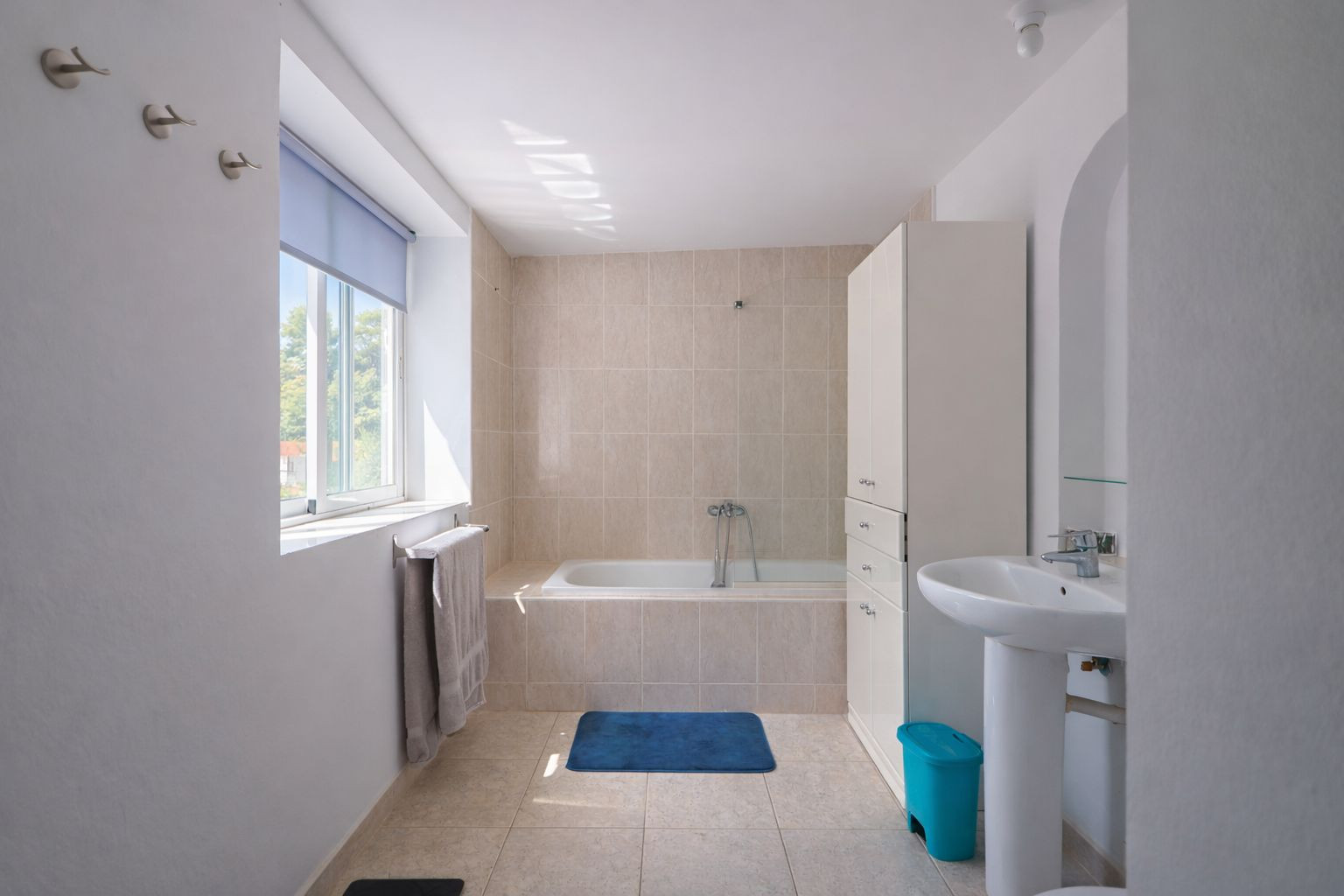 Functional bathroom featuring a built-in bathtub, neutral ceramic tiling, a pedestal sink, and a window for natural ventilation.