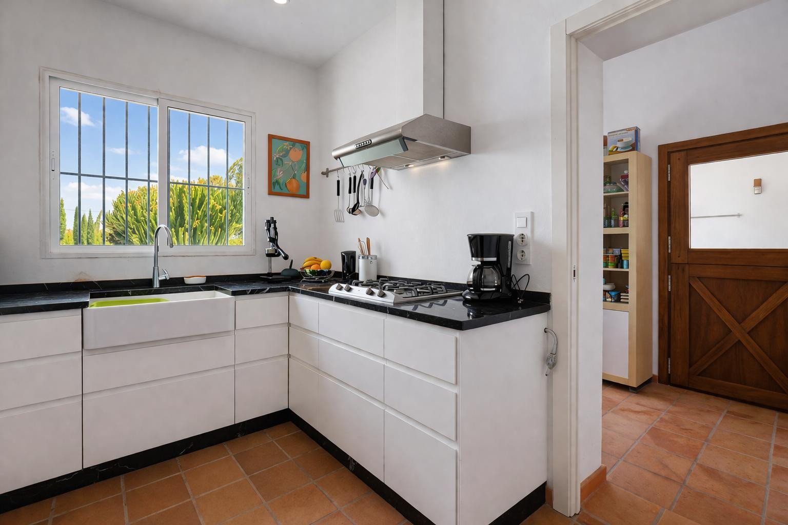 Functional kitchen with black stone countertops, white handleless cabinetry, and a farmhouse sink. Terracotta flooring and natural light define this bright workspace.