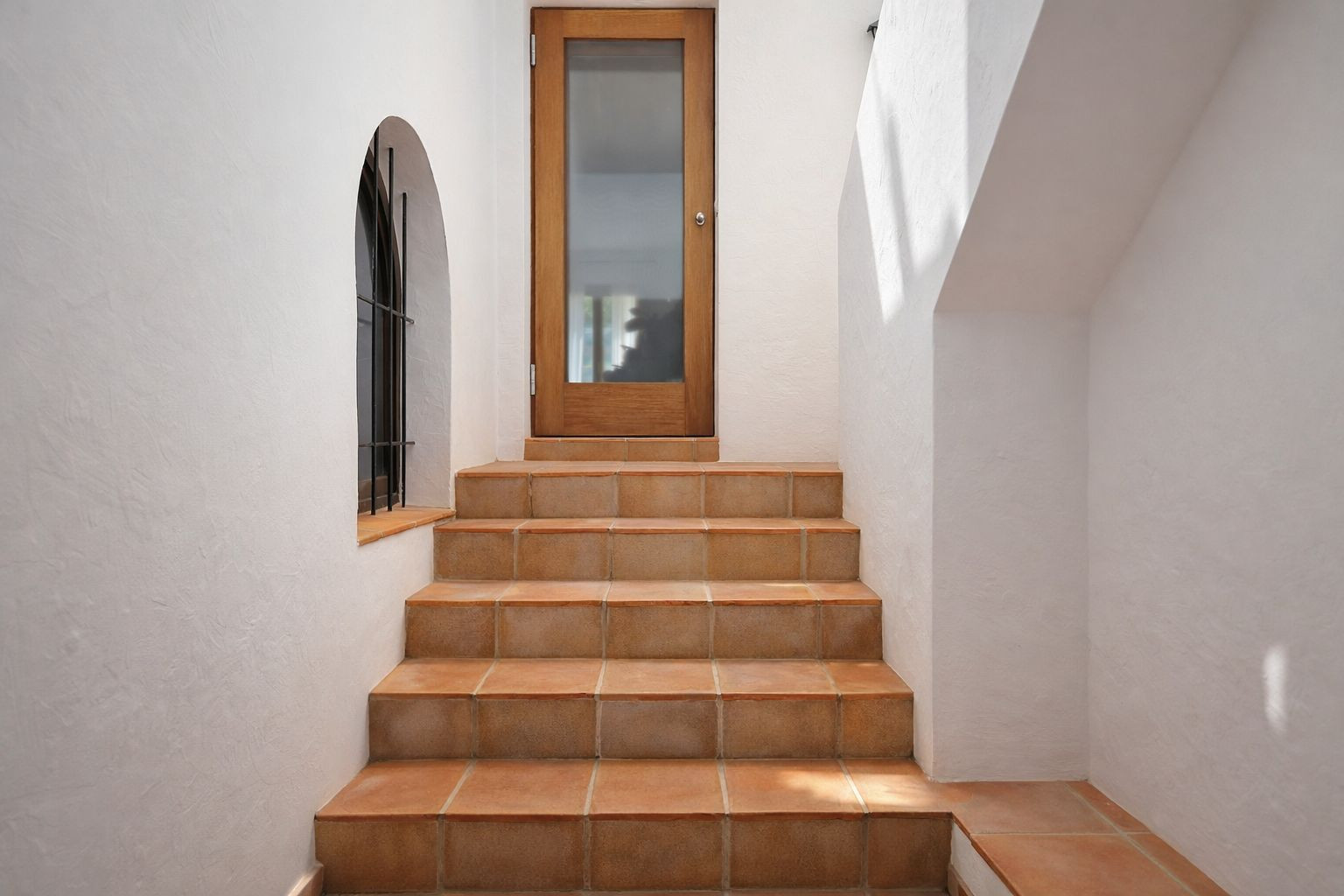 Interior staircase with terracotta tiling leading to a glass-paneled wooden door, featuring a traditional arched window with wrought iron detailing.