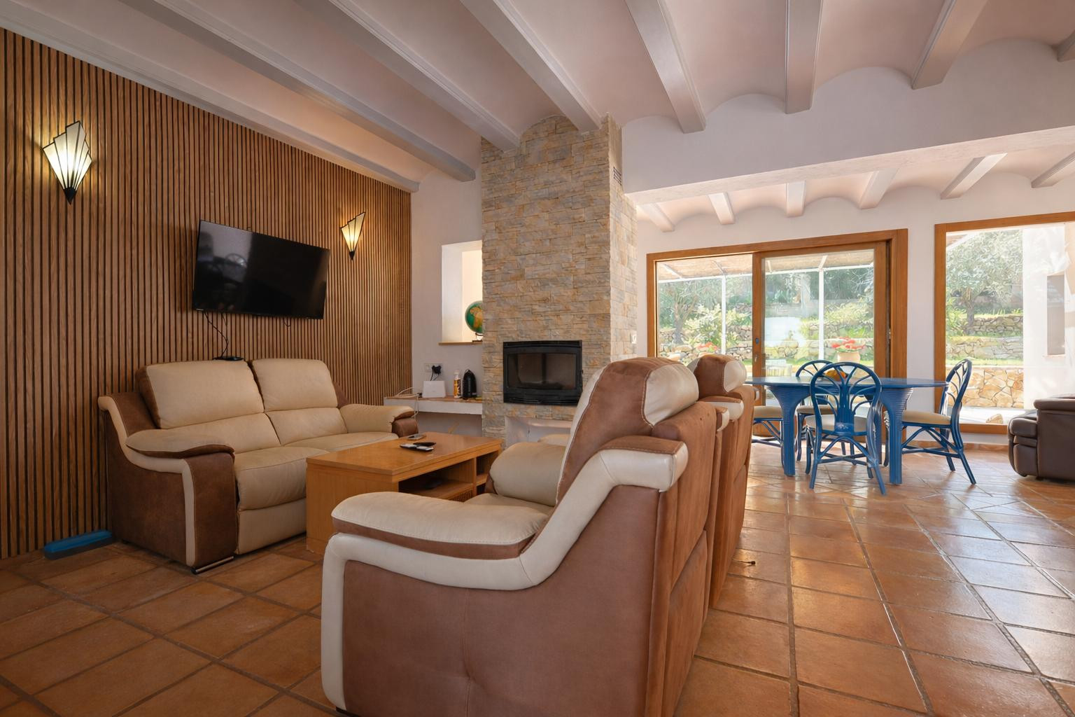 Living and dining area featuring a stone fireplace, white beamed ceilings, and terracotta flooring. Includes a wood-slatted accent wall and sliding glass doors to the porch.