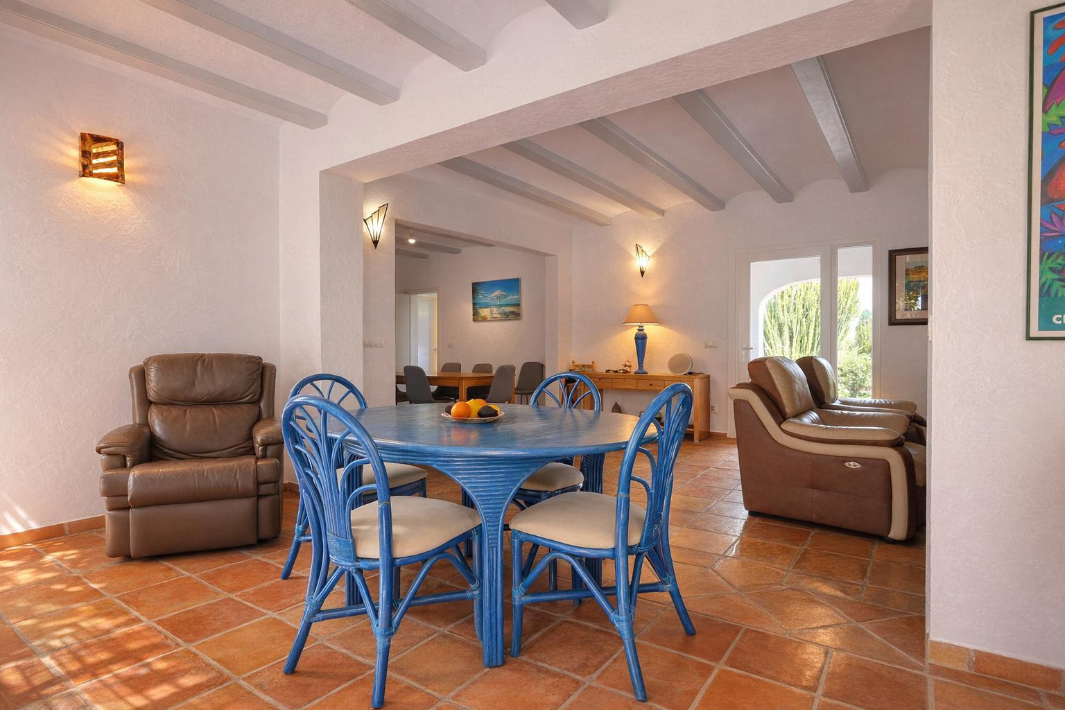 Living-dining area featuring terracotta tile floors, exposed white ceiling beams, and blue rattan furniture. Open archways lead to a secondary lounge and dining space.