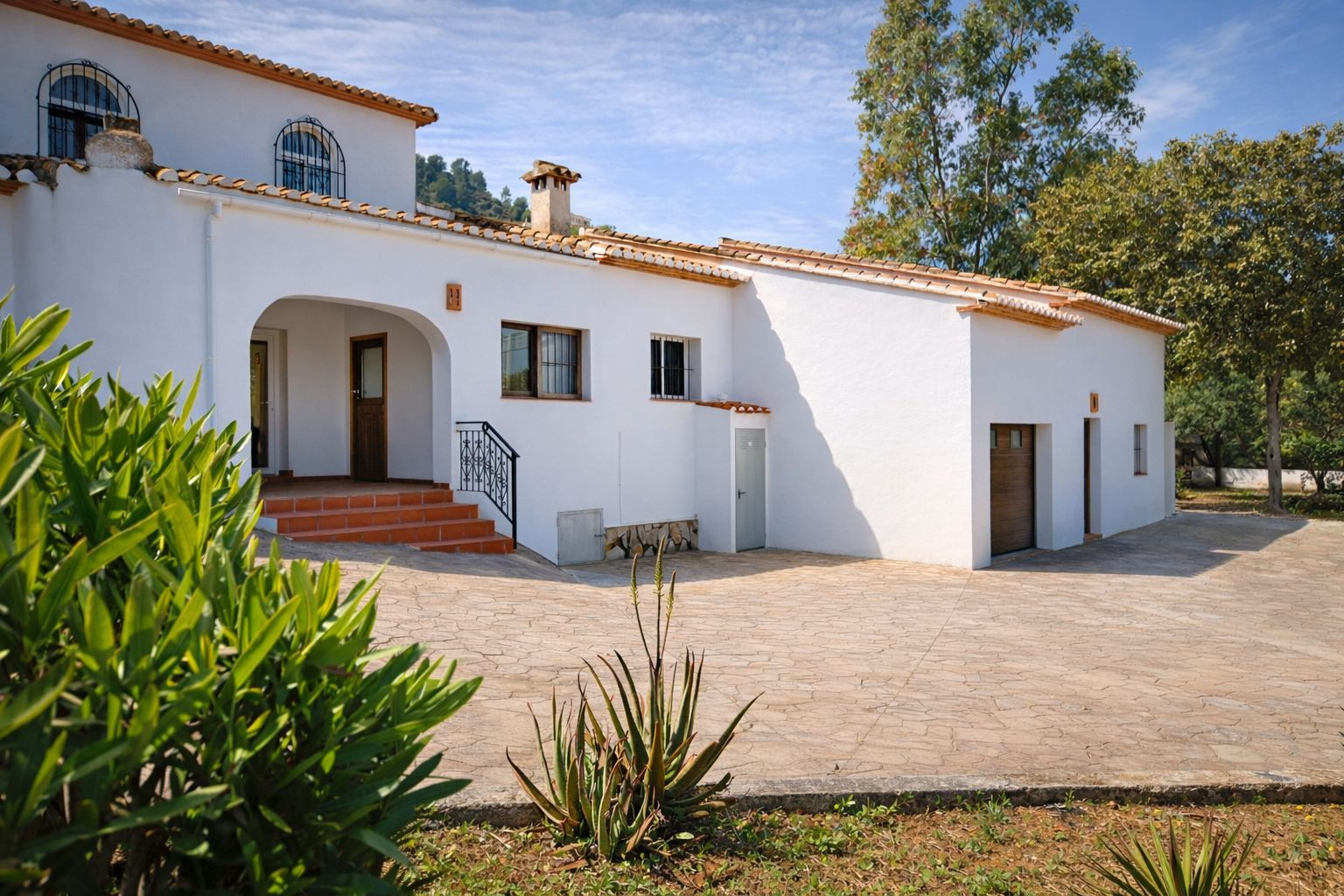 Mediterranean-style white facade featuring terracotta roof tiles, an arched entrance porch, and a spacious stone-paved driveway leading to the integrated garage.