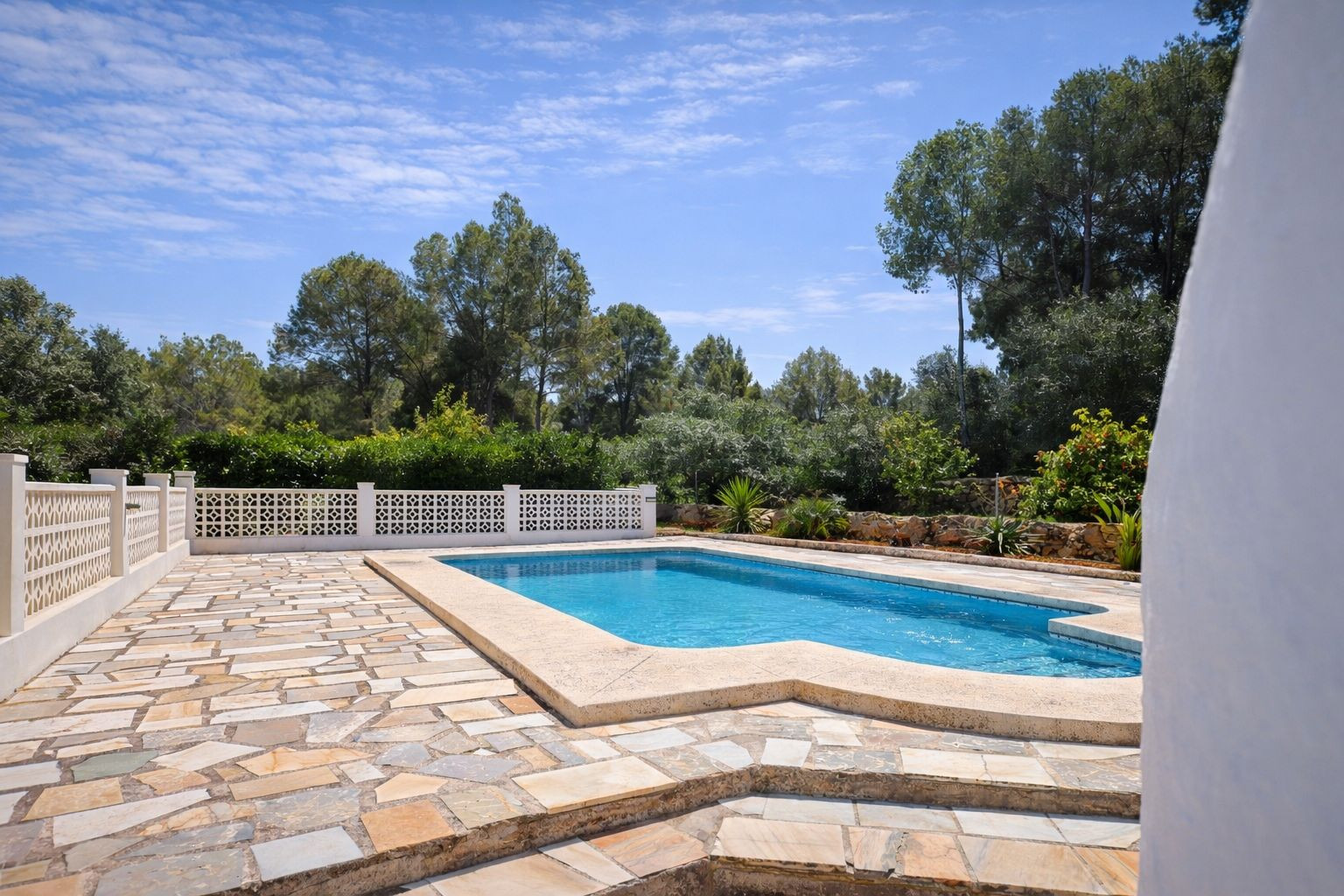 Outdoor swimming pool featuring a natural stone terrace, decorative white lattice fencing, and a backdrop of mature Mediterranean trees.