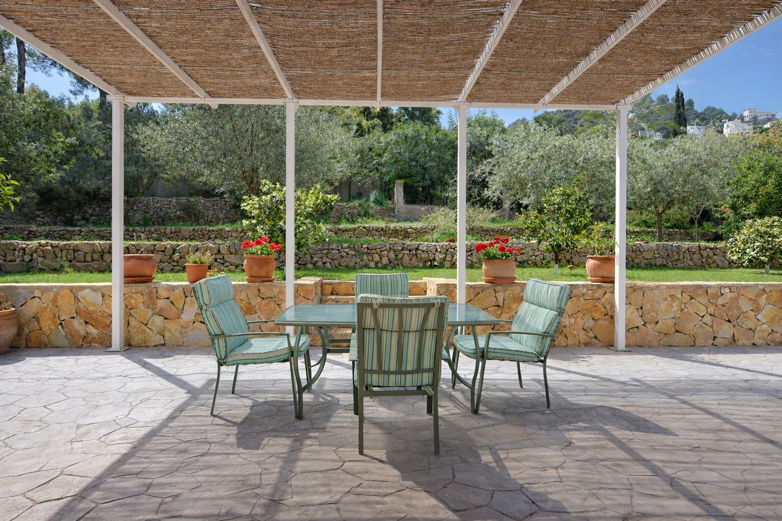 Outdoor terrace featuring a reed pergola, dining set, and views of a terraced Mediterranean garden with natural stone walls and fruit trees.
