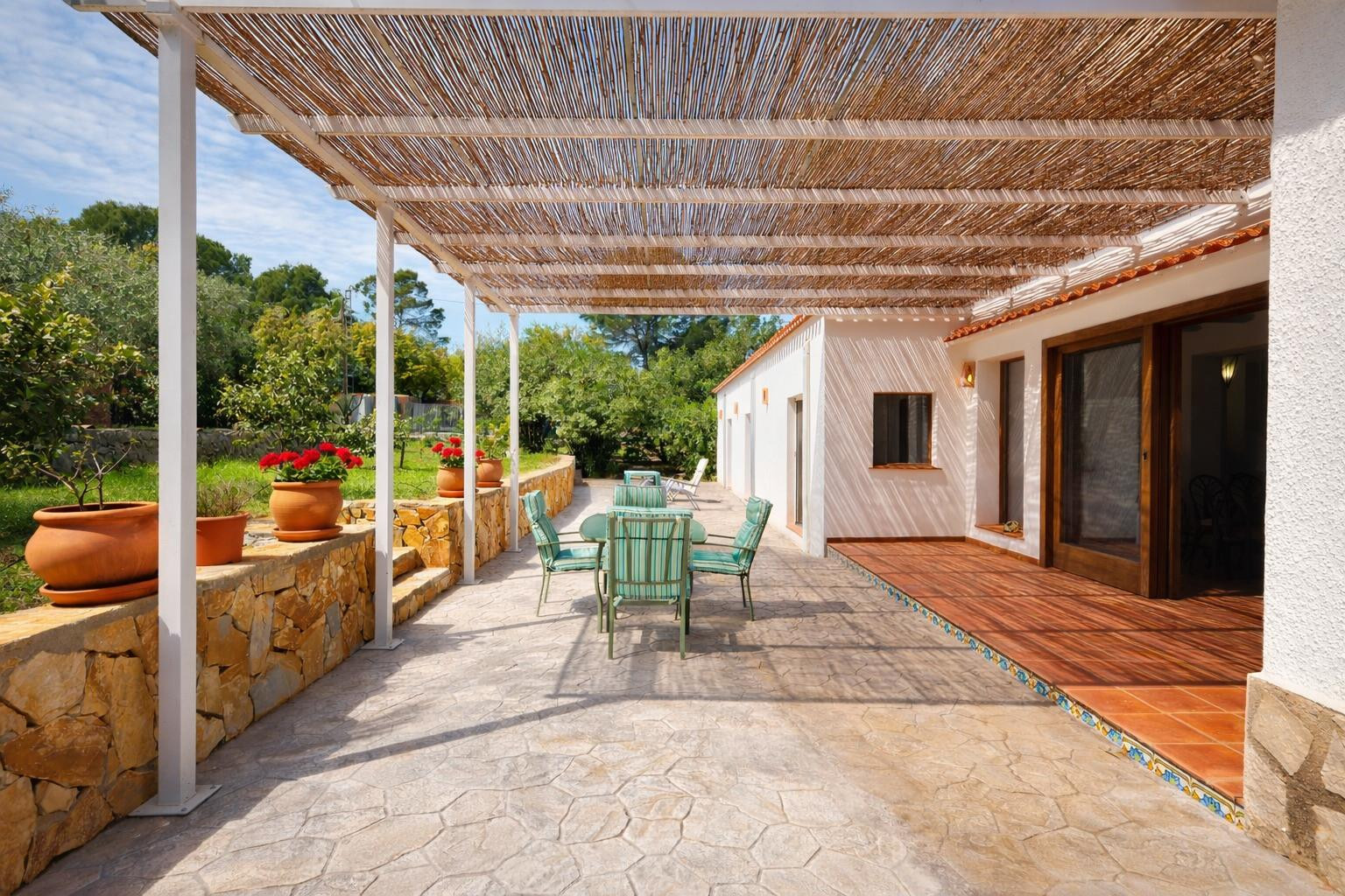 Outdoor terrace featuring a reed pergola, stone-paved flooring, and a natural stone wall with terracotta planters. The space includes a dining area and garden views.